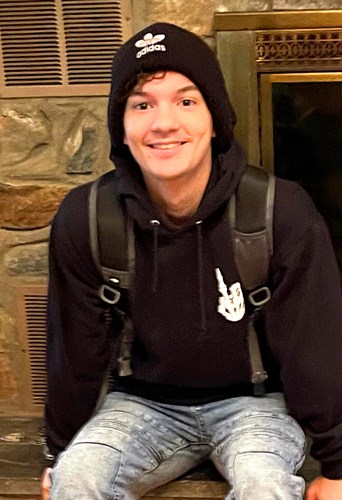 A young boy in a black hooded sweatshirt, blue jeans and black beanie is smiling at the camera.