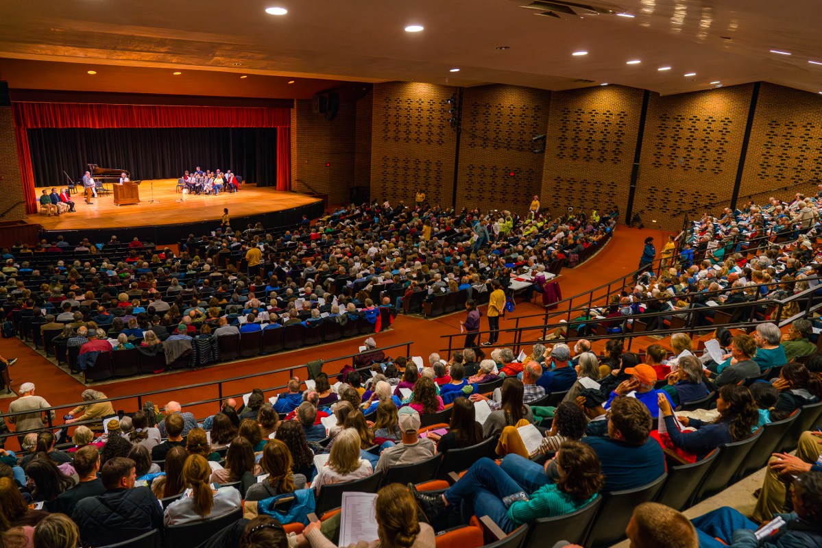 A long view of an indoor auditorium with many rows of seats. Most of the seats are full. In the distance, there are people sitting in chairs and standing on a stage.