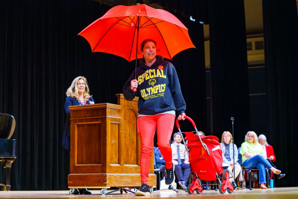 A woman walks across an auditorium stage, holding an open umbrella in one hand and pulling a wheeled backpack behind her. She is wearing a sweatshirt that says "Special Olympics" across the chest. Another woman stands a few feet away, behind a podium, watching her. In the distance, a few people are seated in chairs, watching the action.