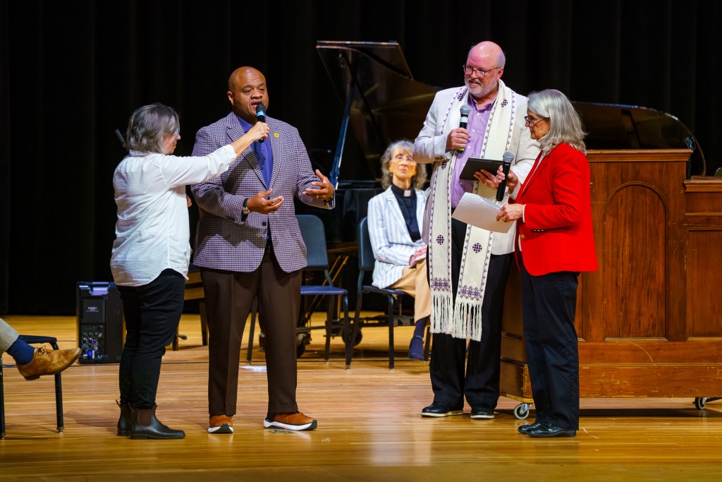 Four people stand on an auditorium stage. A woman on the left holds a microphone up to a man who is speaking and gesturing with his arms. Two people stand to the right, also holding microphones, watching the man who is talking.
