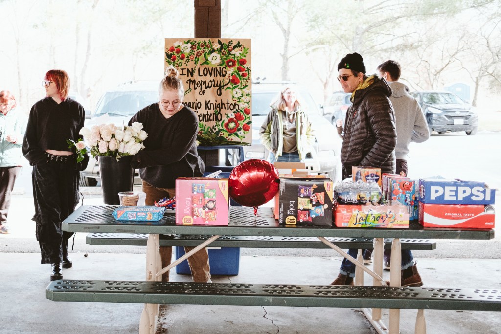 A young woman is setting a bucket with white roses on a picnic table filled with snacks and drinks. Several people walk or stand around her, and a hand-painted sign behind her reads, "In loving memory of Demario Knighton Centeno."