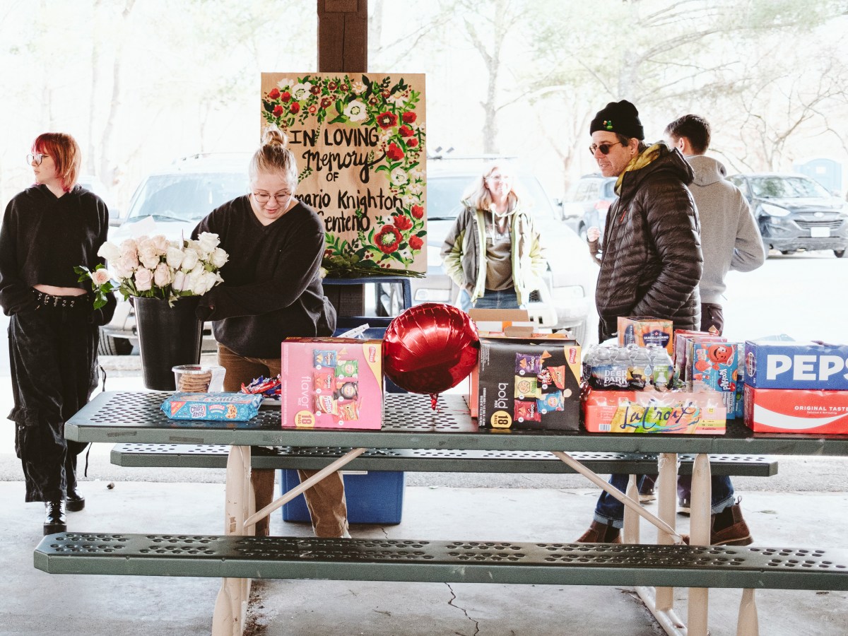 A young woman is setting a bucket with white roses on a picnic table filled with snacks and drinks. Several people walk or stand around her, and a hand-painted sign behind her reads, "In loving memory of Demario Knighton Centeno."