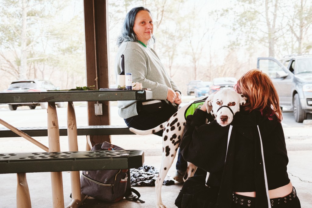 A woman is sitting at a picnic table and looking at the camera. On the ground, another person is sitting back to the camera and hugging a dalmatian in a bright green vest.