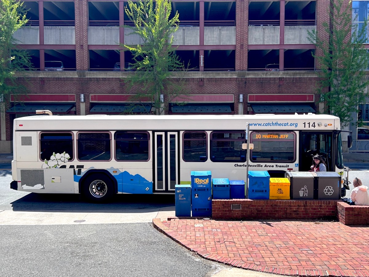 A city streetscape. In the center of the photo, a public transit bus is pulled up to the curb to let passengers off and on. There are newspaper boxes, a trash can and a recycling can in the foreground in front of the bus, and a multi-story parking deck in the distance behind it. A digital screen near the front door of the bus says "10 Martha Jeff" to indicate the route it is running.