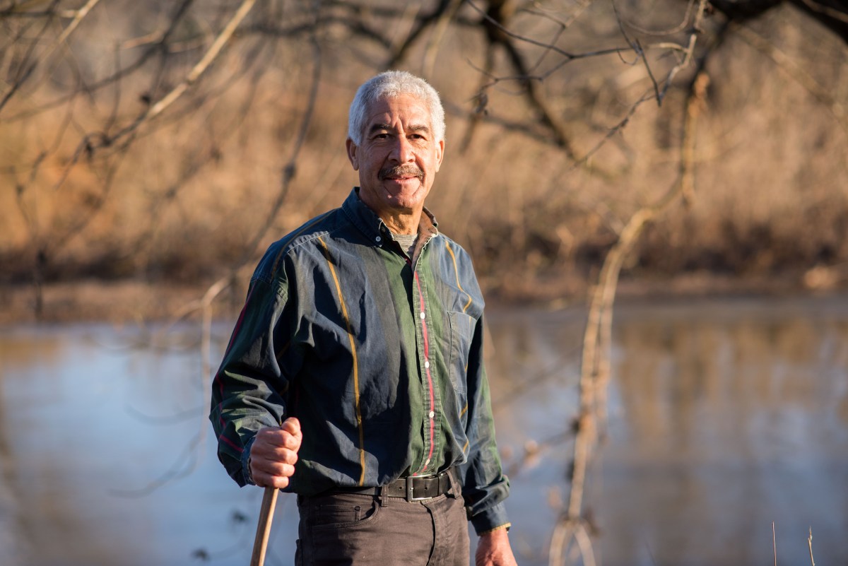 A man with a walking stick looking at the camera, a fall river scene behind him.