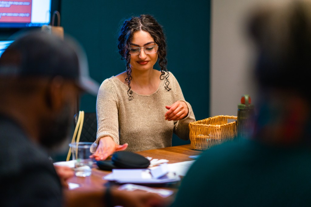 A woman sits at a table working with others who are out of focus around her.