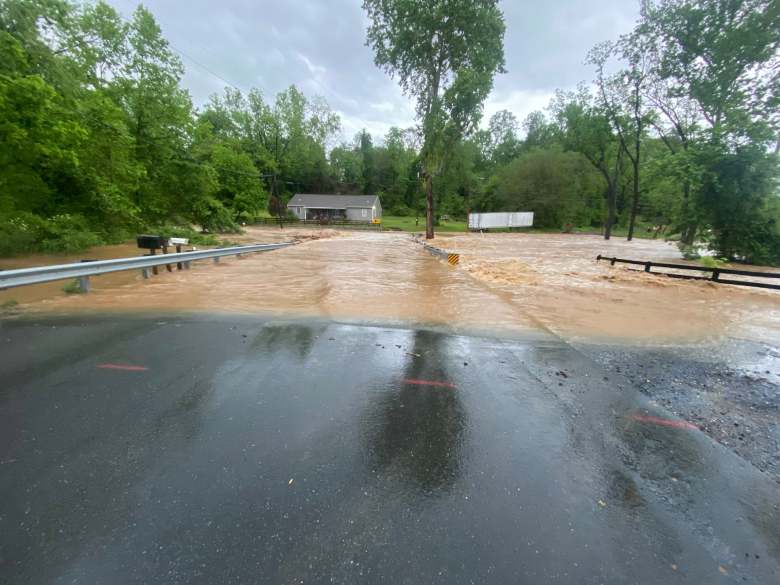 A road with trees, a barn and a trailer in the background. Rising water covers the road and is rushing over the side of the road, nearly submerging a nearby fence.
