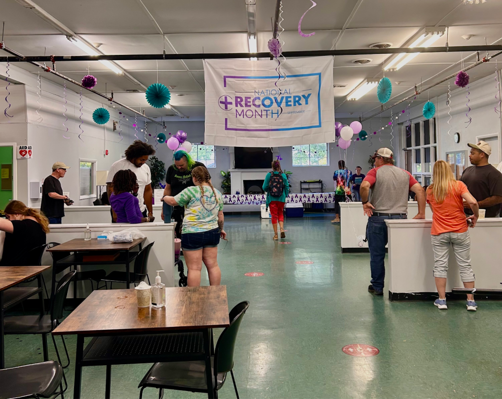 Groups of people stand in a large room with mostly empty tables and chairs spread around the area. Paper decorations and a banner saying "National Recovery Month," hang from the ceiling.