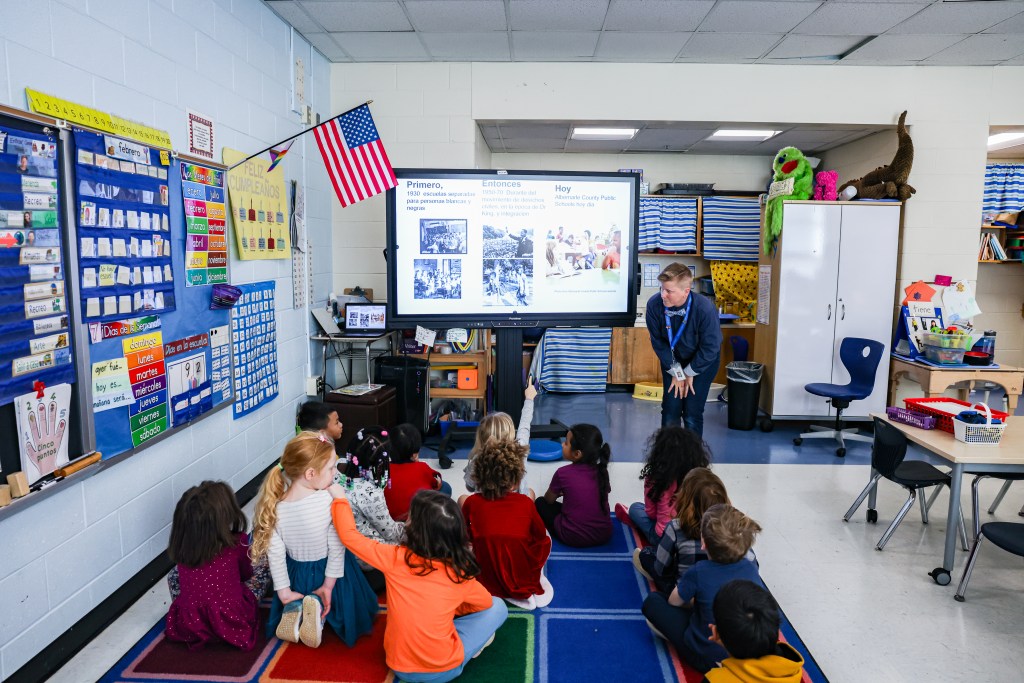 Several small children are seated on the floor of a classroom with chairs, an American flag and several posters on the wall. They look toward a woman standing at the front of the room next to a screen with a presentation on it.