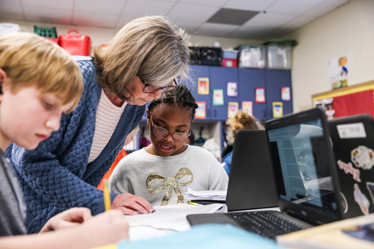 A woman wearing glasses leans over a desk. looking at a paper with a student. Another student sitting next to her is writing something down on another piece of paper. Both students have laptop computers open in front of them on the desk.