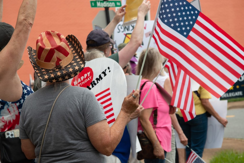 People are gathered next to the road holding large American flags and signs supporting the Army. One of them is wearing a red white and blue cowboy hat.