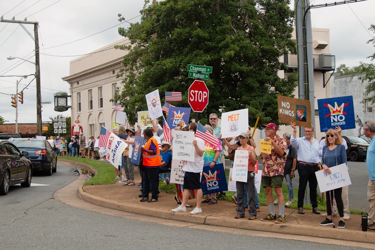A public street with several people holding colorful signs, many reading 'No Kings," and holding American flags while standing on the sidewalk. Two cars are passing by on the road.