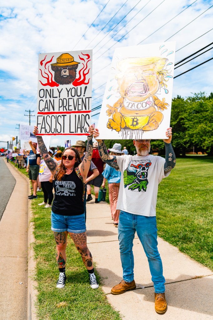 Two people stand on a sidewalk next to a road holding up large homemade signs, one depicting President Donald Trump as a crying baby and the other reading "Only you can prevent fascist liars." Several dozen other protestors are standing on the sidewalk behind them.