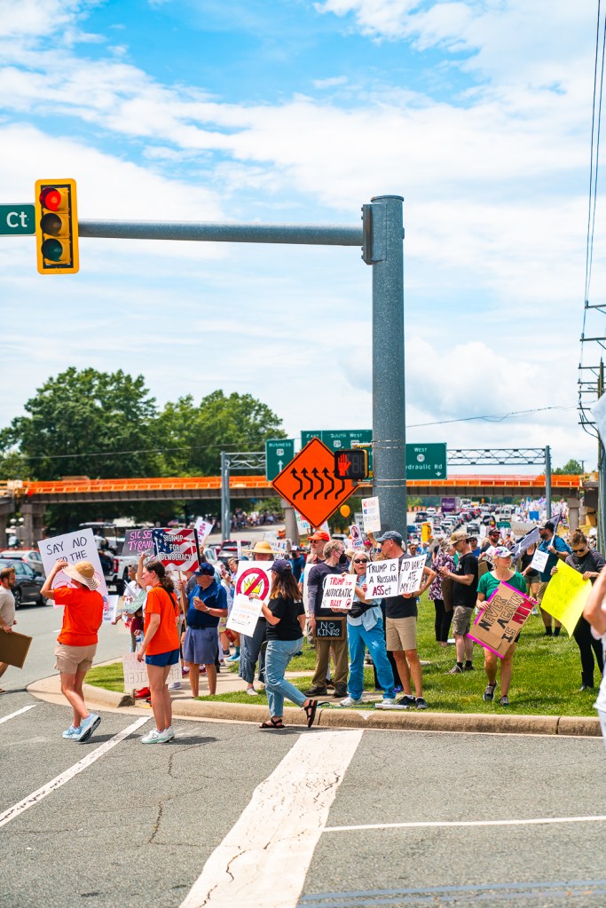 A large group of people stand at an intersection with a traffic signal and a large orange sign indicating road work.