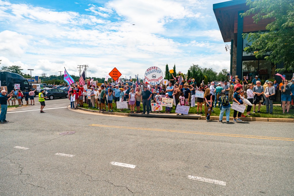 A large group of people gather on a sidewalk holding up colorful signs and American flags. A person in a reflective vest stands off to the side directing traffic as cars drive by.