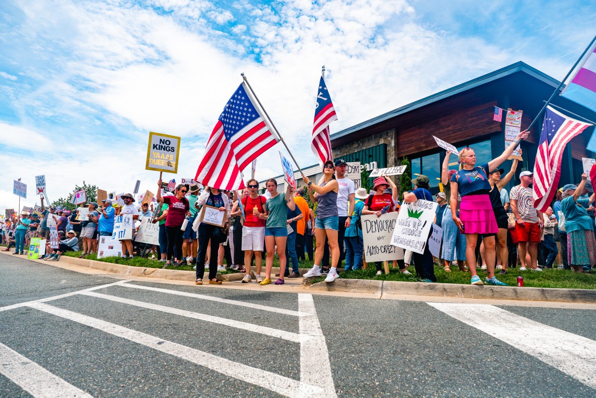 A large crowd of people gathered on a sidewalk next to a road, waving large American flags and holding protest signs.