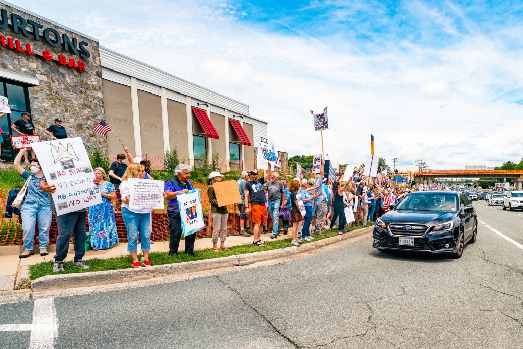 Hundreds of people stand on a sidewalk next to the road holding up protest signs as cars pass by.