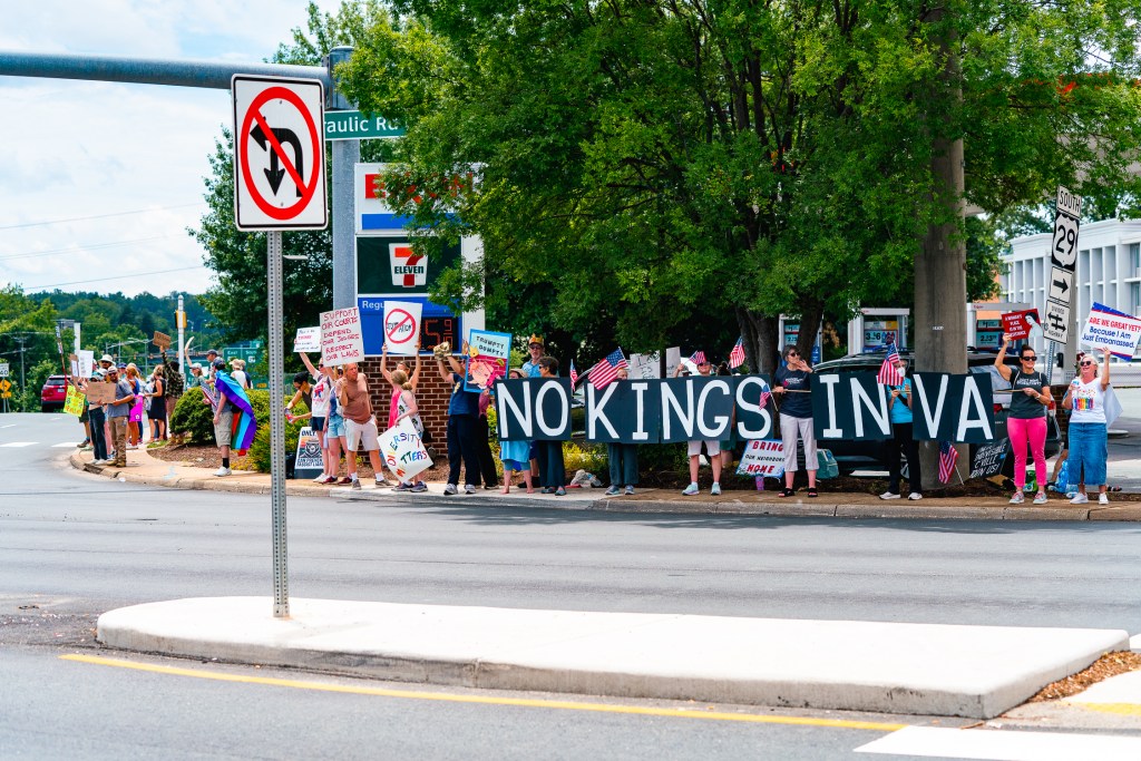 Several people stand on a street corner in front of a gas station holding up protest signs, the largest of which reads "No kings in VA."