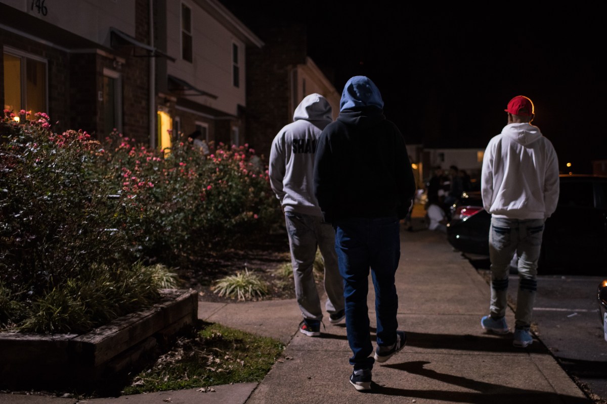 Three men in hoodies walk along a sidewalk at night next to two-story town houses.