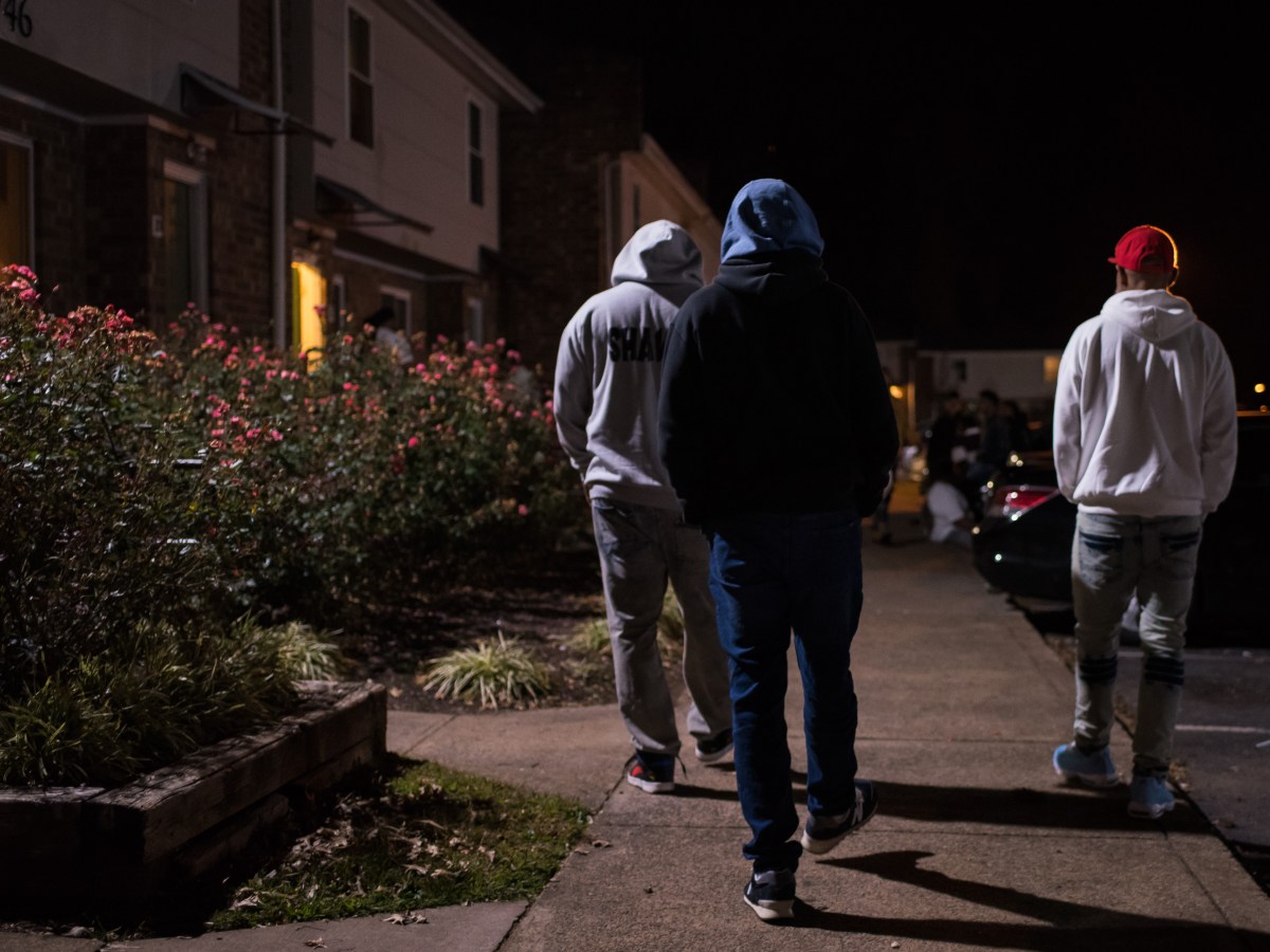 Three men in hoodies walk along a sidewalk at night next to two-story town houses.