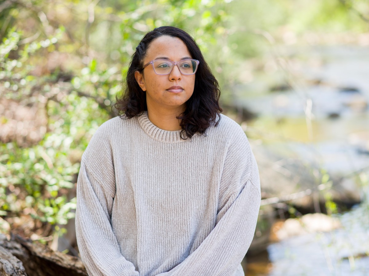 A person with shoulder-length dark hair and glasses looking up to the sky, sun falling on her face, in front of greenery.
