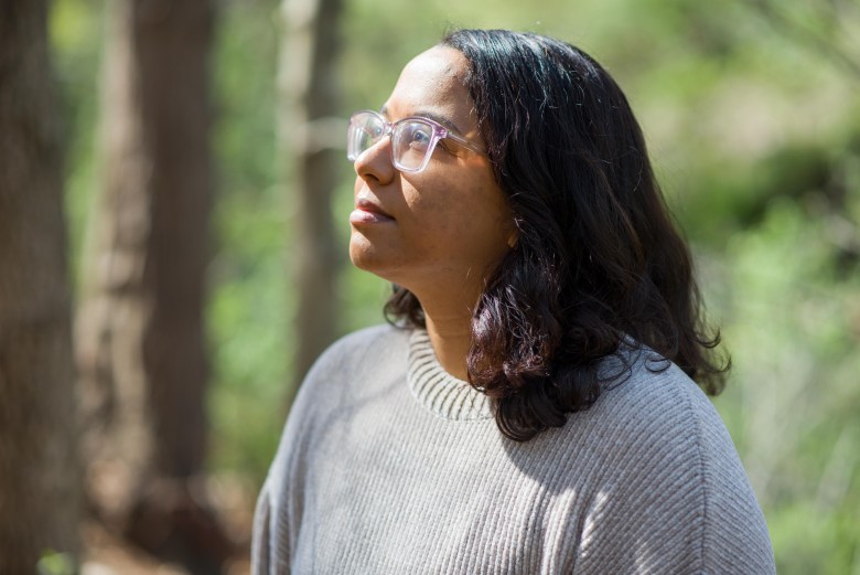 A person with shoulder-length dark hair and glasses looking up to the sky, sun falling on her face, in front of greenery.