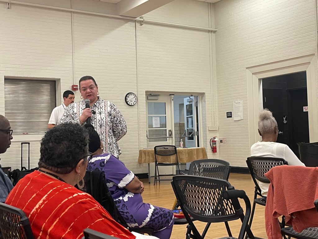 A woman stands in a room with wooden floors and painted white brick walls speaking into a microphone. People sit in folding chairs near her, listening, and other people stand against the wall behind her.