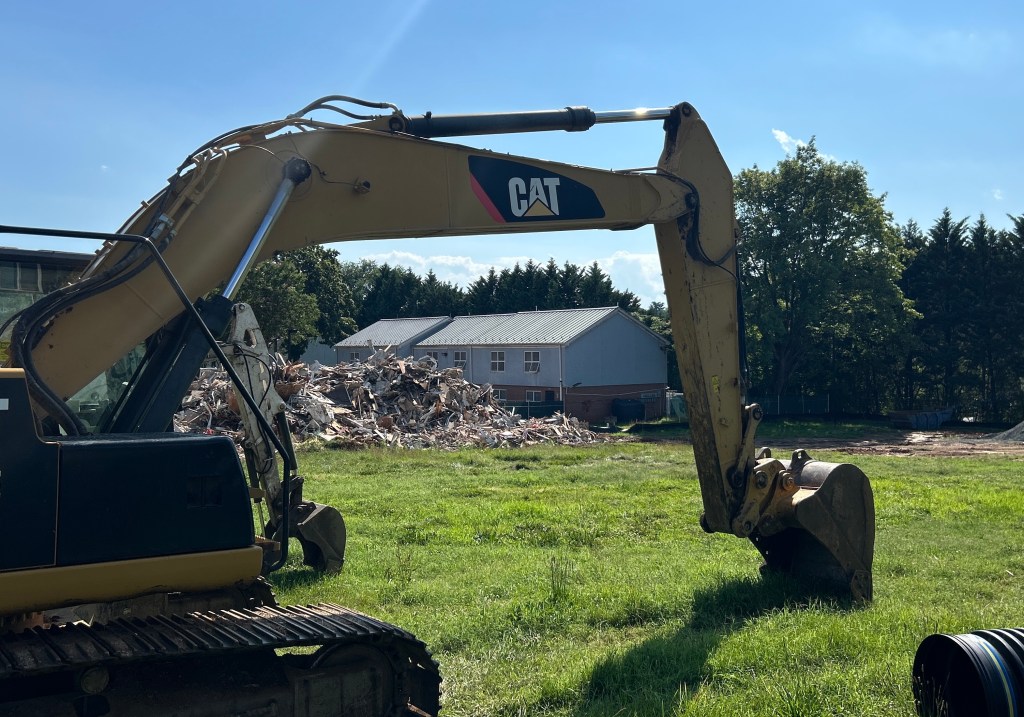 An excavator stands still on some grass on a sunny day. The arm of the excavator makes a frame for a row of two-story townhomes and a pile of debris from a recent demolition.