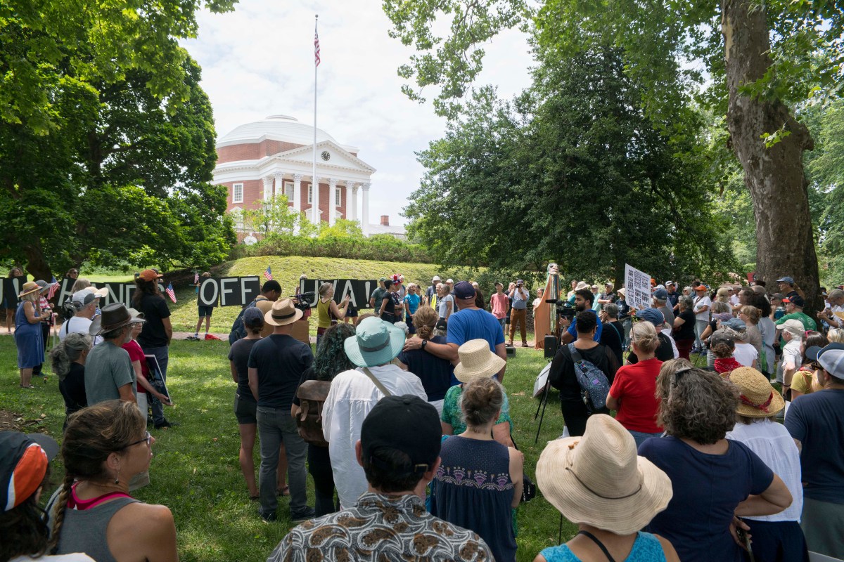 A crowd of people holding signs gather on the grass near the rotunda, a large brick building with white columns. Several people hold up a large sign reading "Hands Off UVA."