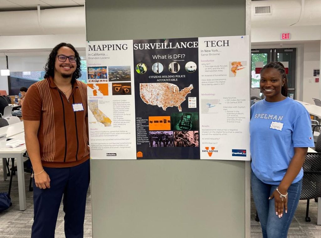 A man and a woman stand indoors next to a large poster titled "Mapping Surveillance Tech" covered with colorful graphics and a map of the United States. Desks, tables and computer monitors are visible in the background.