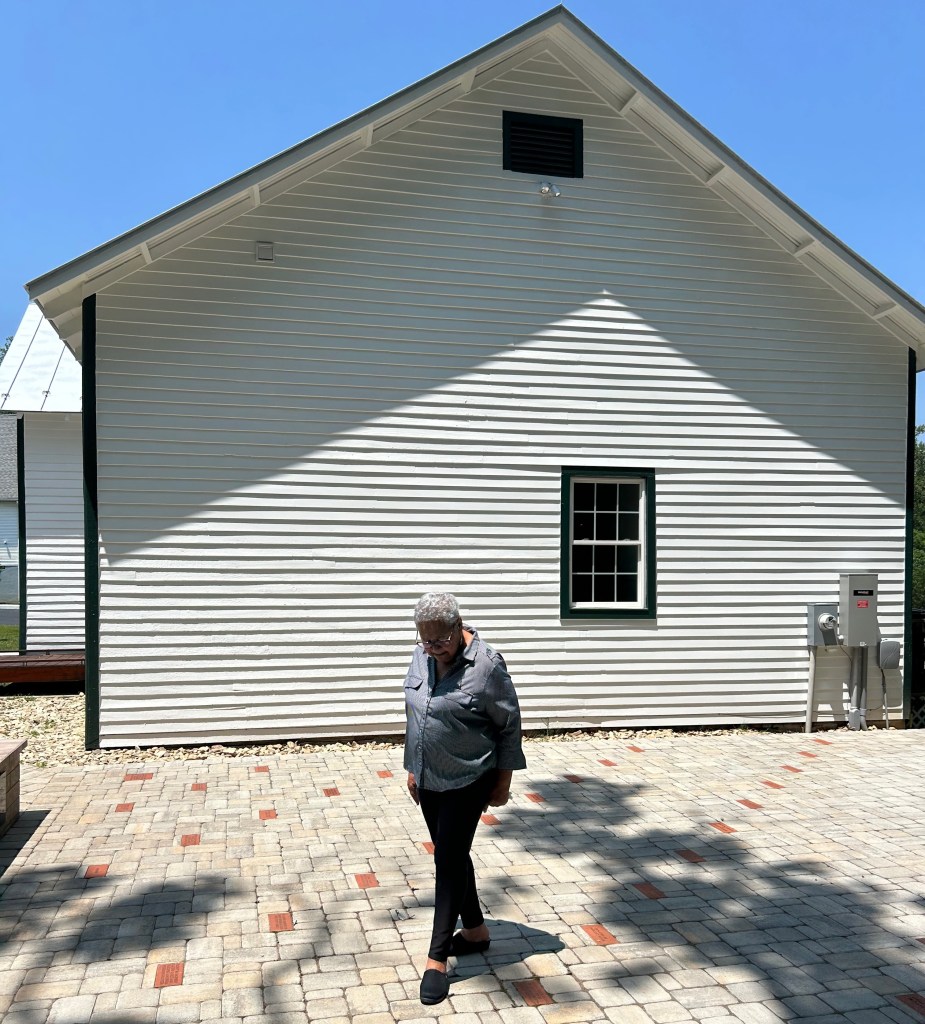 A woman stands outside, next to a white building with black trim, looking down at a cobblestone and brick courtyard. Shadows of trees are visible on the ground.