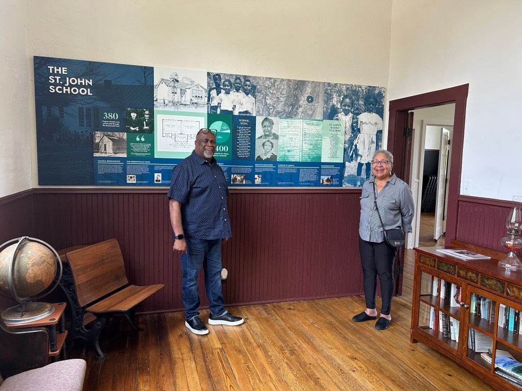 A man and a woman stand in a large room with a bookcase, chairs and a large globe. Behind them is a sign reading "The St. John School" with several black-and-white pictures and text.