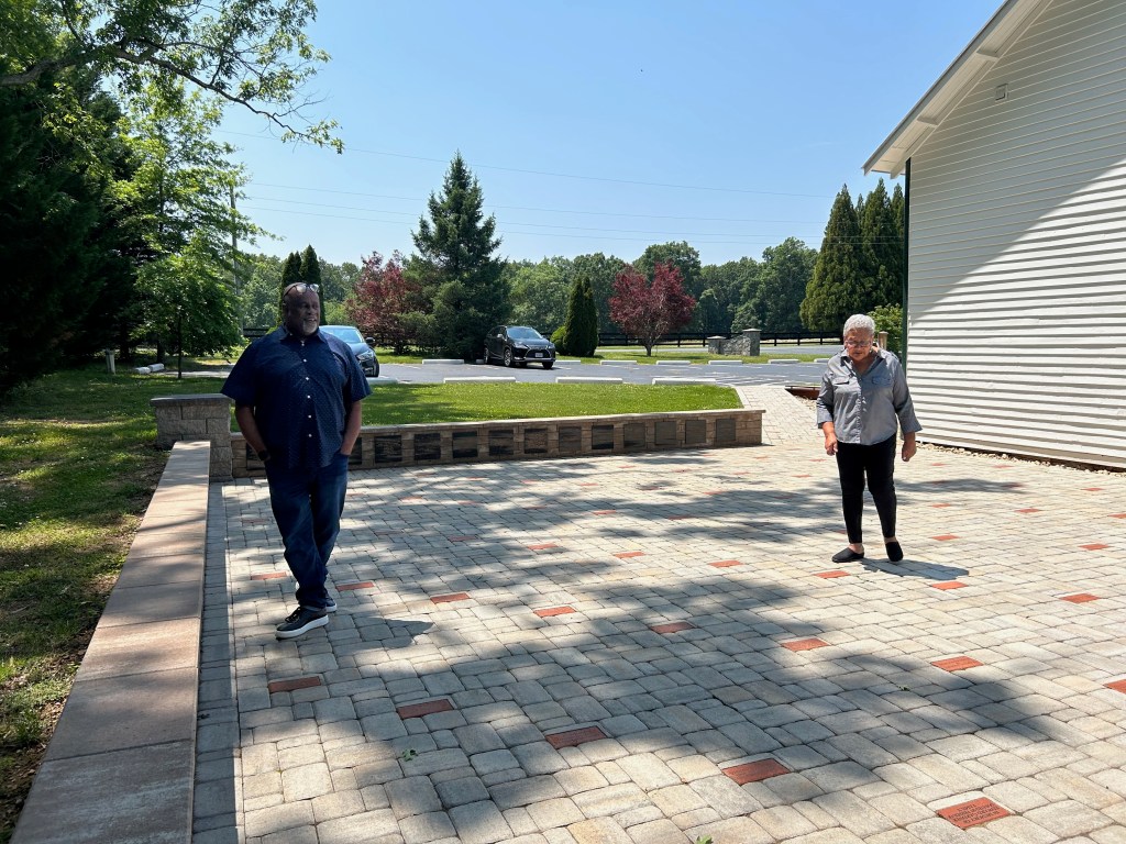A man and a woman stand on a stone and brick patio, with several bricks and plaques inscribed with the names of donors or community members. Grass, trees and a parking lot are visible behind him.