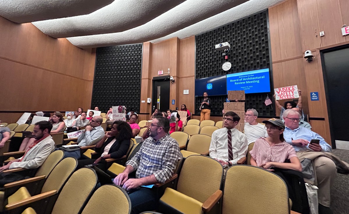 People sit in rows of chairs in a small auditorium with wood paneling on the walls behind them. They look toward the front of the room, and many of them are holding up homemade signs, one reading "Justice, Safety, Access."