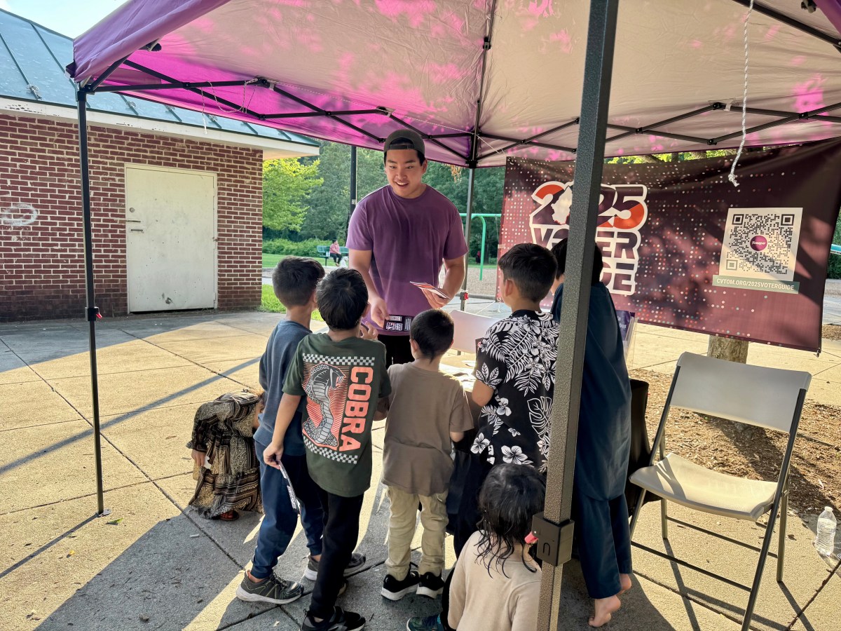 A young man hands out flyers to children at a tabling event, under a purple tent. In the background a large banner reads "Voter Guide 2025."