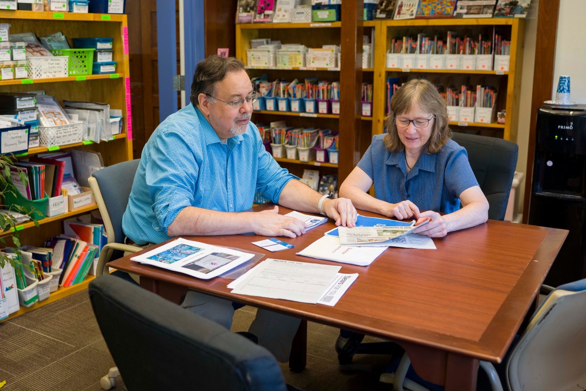 wo people sit at a table looking over stacks of paper. Behind them are bookshelves full of different-sized containers holding books, flashcards, and other learning materials. Each container is labeled with what it holds.