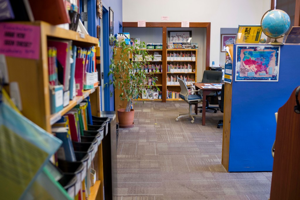 A view of an office. There are many bookshelves packed with books, posters of the United States, a globe, some plants, and tables for folks to sit at during tutoring sessions.