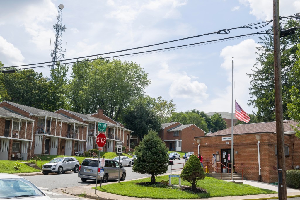 A community of two-story brick townhomes with porches, taken in the summer. There is a radio tower in the background, as well as a glimpse of a tall apartment building. In the near distance, two people stand talking outside a community center.