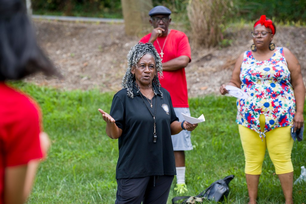 People standing in a park. A woman stands at the center of the photo, speaking. Her arms are slightly outstretched, and she holds a piece of paper in her left hand. Behind her, a man and a woman listen closely to what she is saying.