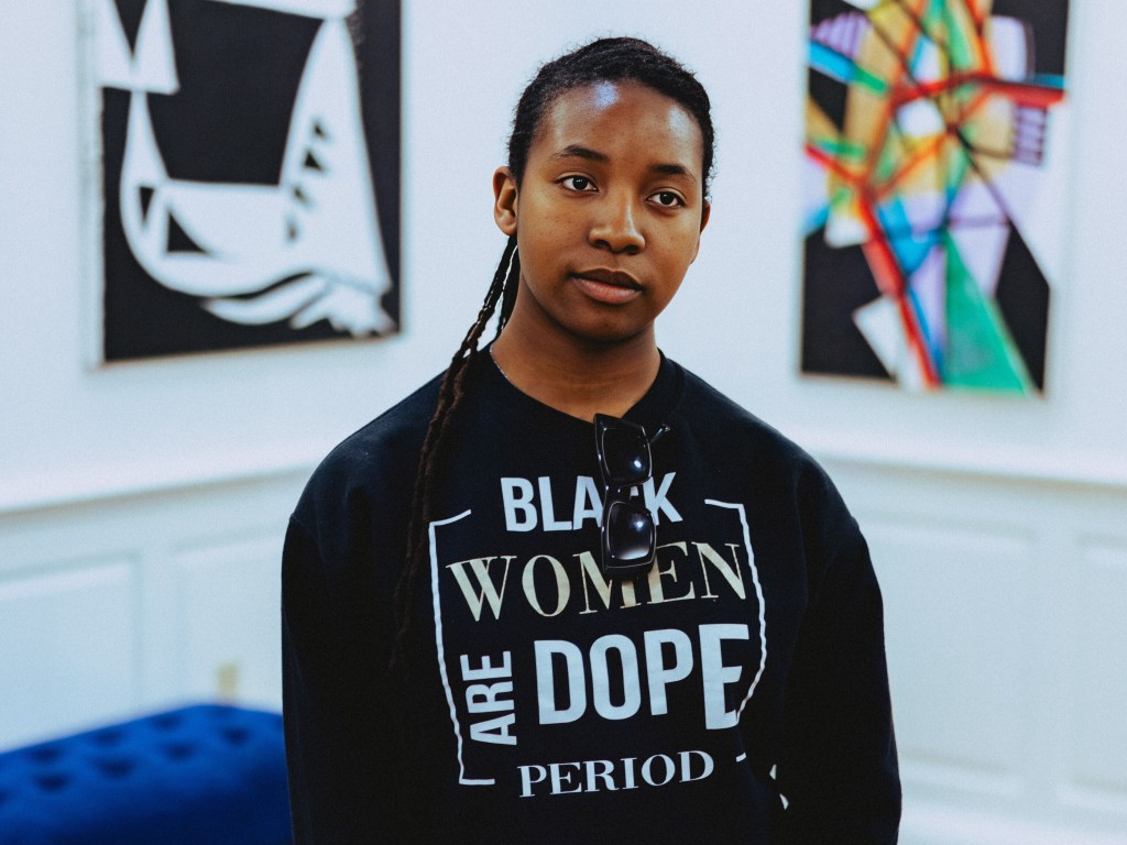 A young woman stands with her arms clasped. Her shirt reads "Black Women Are Dope Period." Abstract art hangs on the wall behind her.