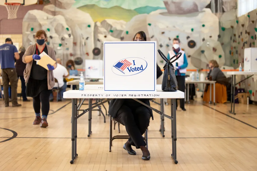 A person sitting at a voting booth in a gymnasium, holding a ballot, with 'I Voted' sticker visible on the booth, while other voters are seen in the background.