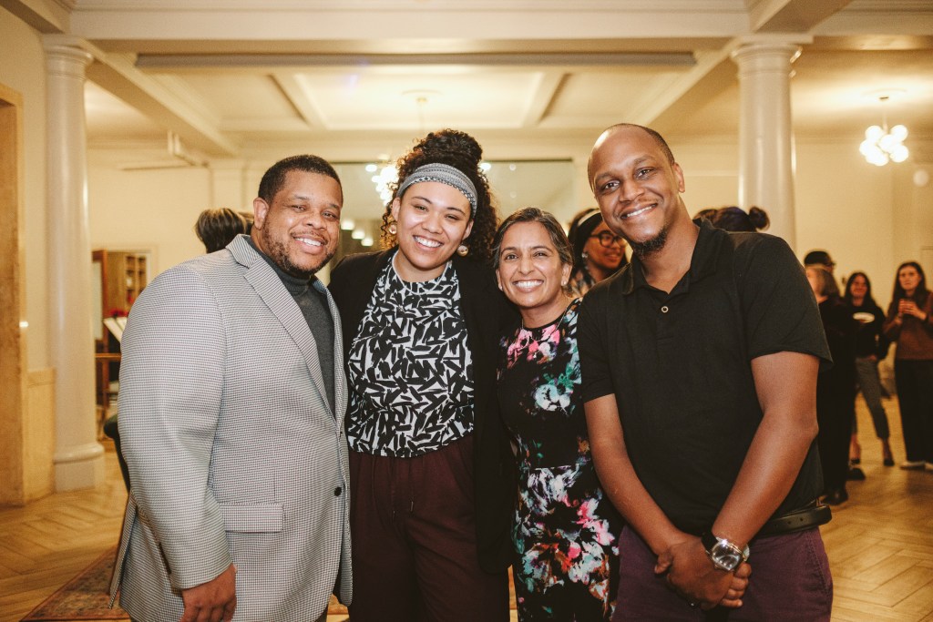 Four people in a glowing room stand together, with more people behind them. Left to right, a man in a gray suit jacket, a woman in a headband with a black and white patterned top, a woman in a floral pattern dress and a man in a black polo shirt.