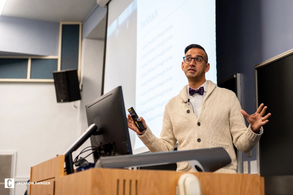 A person in a bowtie and sweater stands at a podium in a college lecture hall, speaking.