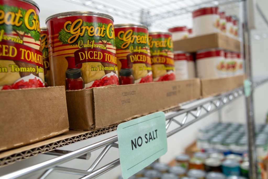 Metal shelves stocked with cans of diced tomatoes and other vegetables. A sign attached to the shelf reads "No salt."