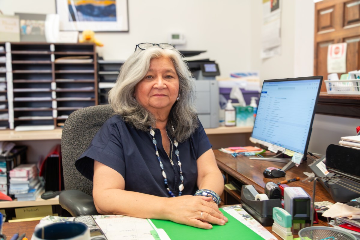 A woman with glasses pushed up on her head sits at a large wooden desk with a computer monitor, folders, a tape dispenser and other items.