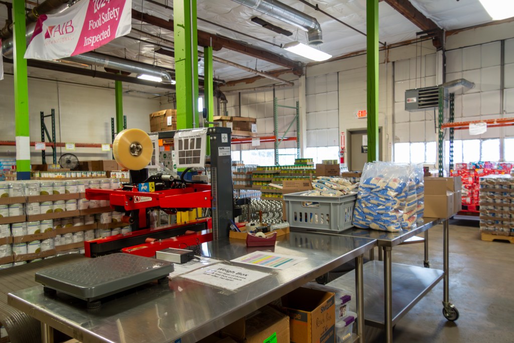 A metal table with a large scale sits in an open warehouse. Stored around the room are pallets and plastic crates stacked with canned vegetables and other non-perishable food.