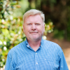 A man with a beard in a blue button-down shirt smiles for camera, greenery behind him.