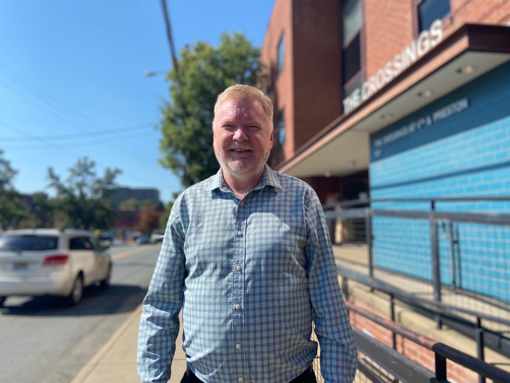 A man in a button-down checker blue shirt smiles, standing on the sidewalk next to a brick and tile building with a sign that says "THE CROSSINGS." A blue sky and white car driving by are blurry in the background.
