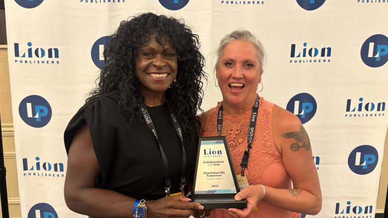 Two women in front of a repeater banner that says "Lion" with an "L" logo hold an award and smile.