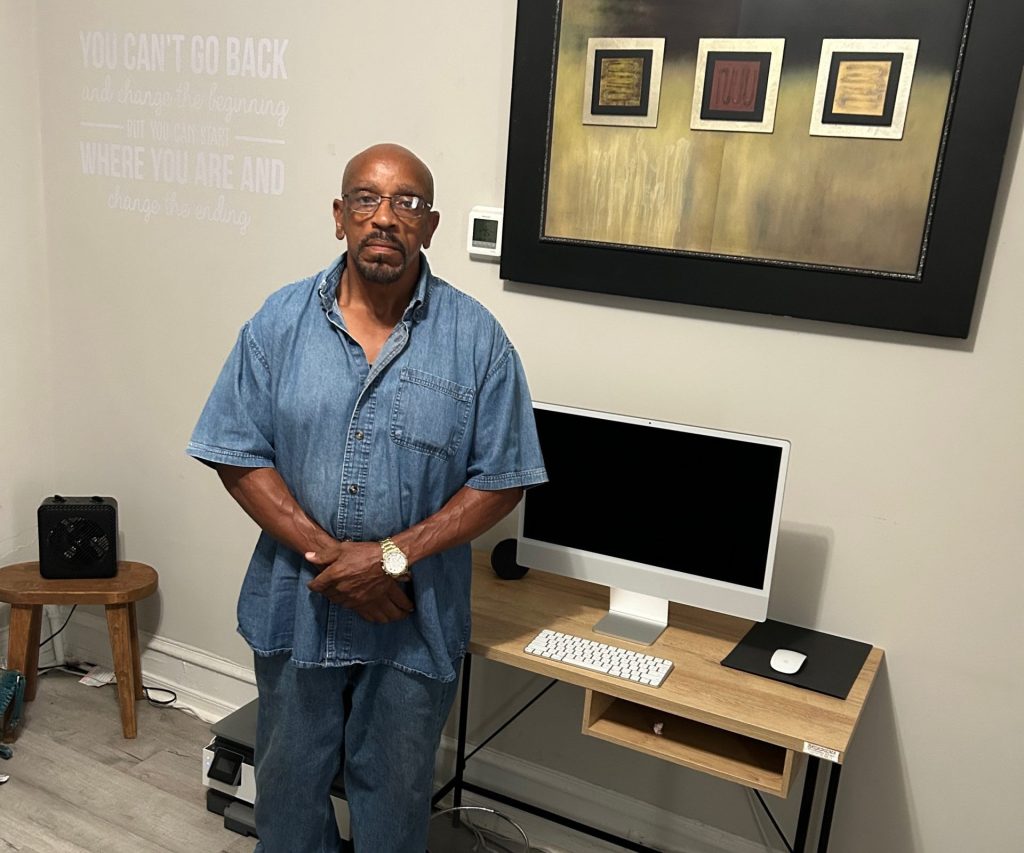 A man wearing a denim shirt and pants stands in front of a desk and computer in a duplex apartment.